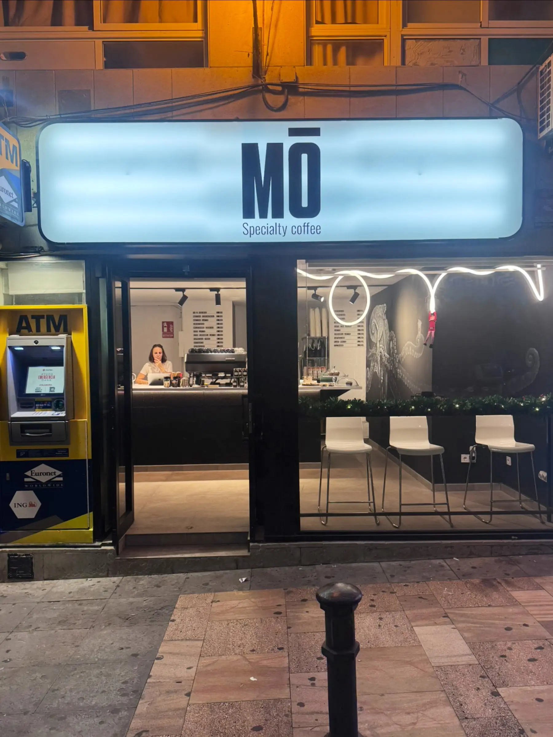 La fachada de MŌ de noche: el rótulo iluminado, la barra y los taburetes blancos en la ventana